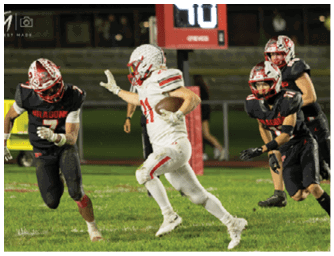 A person in a white football uniform is running with the ball while three people in black uniforms with red helmets are attempting to catch them on a grassy field at night. Descripci n generada con IA