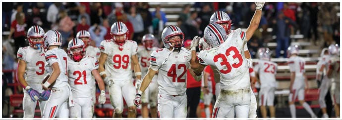 A group of people in white football uniforms with red and black accents are celebrating on a green field during a game. Descripci n generada con IA