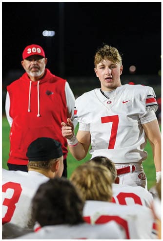 A person in a white sports uniform with red accents stands on a field while another person in casual clothing takes a photo in the background. Descripci n generada con IA
