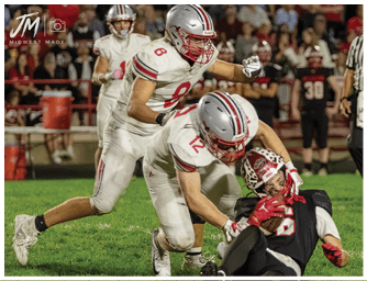 Two people in football uniforms are engaged in a tackle on a grassy field during a game, with one person holding the ball while the other reaches to stop them. Descripci n generada con IA