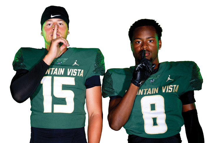 Two adults wearing green football uniforms with numbers 15 and 8 are standing side by side making a shushing gesture with their index fingers to their lips. Description generated by AI
