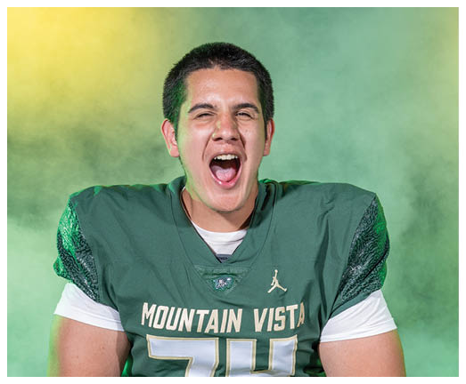 A person wearing a green Mountain Vista football jersey with the number 74 is standing in front of a smoky background with yellow and green lighting, expressing excitement with an open mouth. Description generated by AI
