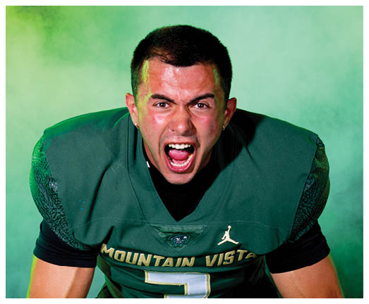 A person wearing a green Mountain Vista football jersey with the number three is leaning forward and shouting with an intense expression against a green smoky background. Description generated by AI