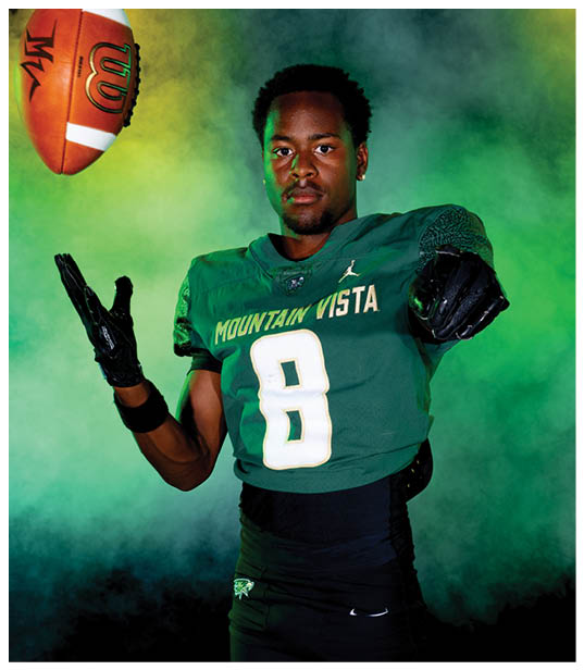 A person wearing a green Mountain Vista football jersey with the number 8 is catching or throwing a football against a green and yellow smoky background. Description generated by AI