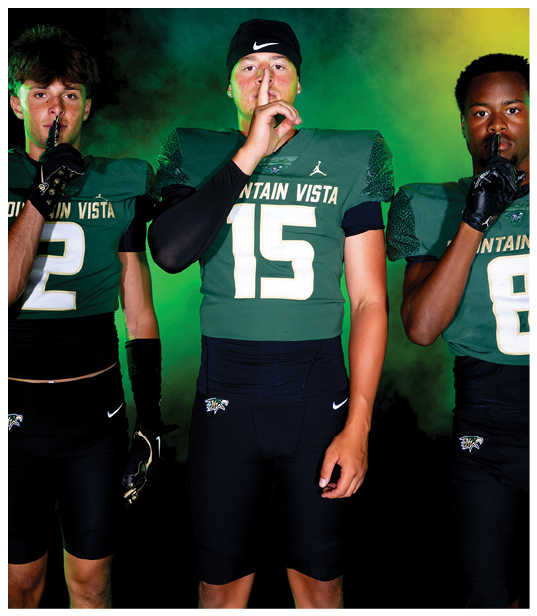 Three people wearing green and black football uniforms with “Mountain Vista" on the jerseys stand side by side holding a finger to their lips in a shushing gesture against a smoky green and yellow background. Description generated by AI