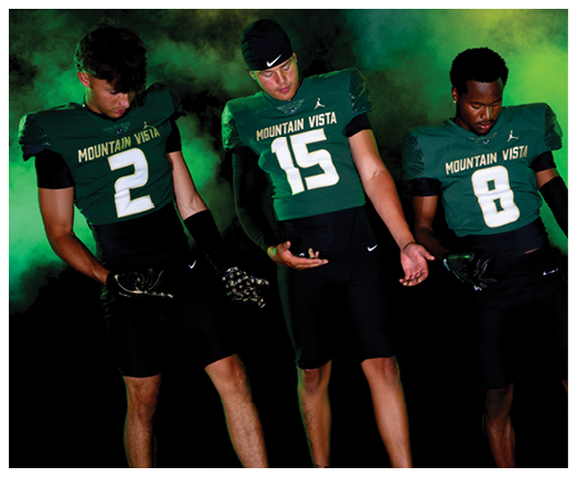 Three people wearing Mountain Vista football uniforms with numbers 2, 15, and 8 stand against a smoky green and yellow background, looking down at their hands. Description generated by AI