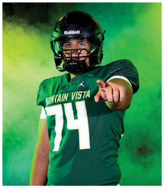 A person wearing a green football jersey with the number 74 and a black helmet is pointing forward against a smoky green and yellow background. Description generated by AI