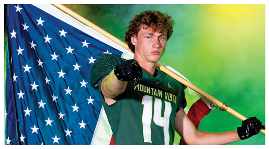 A person with curly hair wearing a Mountain Vista sports jersey points forward while holding a large American flag against a colorful green and yellow background. Description generated by AI