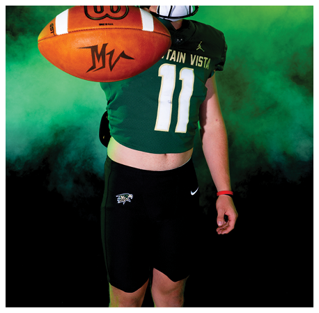 A person wearing a green football jersey with the number 11 and a black helmet is holding a football towards the camera against a smoky green and yellow background. Description generated by AI