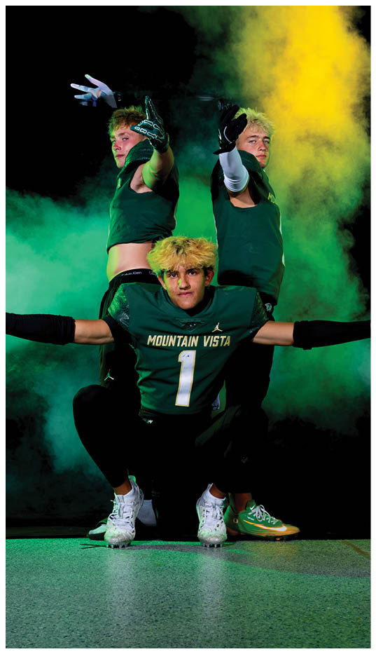 Three adults with blond hair wearing dark green athletic clothing and gloves pose dramatically against a smoky background with green and yellow lighting. Description generated by AI