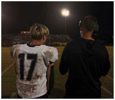 A person wearing a headset stands next to a person in a dirty sports jersey with the number 17 on the back under bright stadium lights at night. Description generated by AI