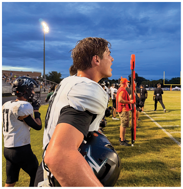 A person with wet hair is shouting or calling out near an orange marker on a sports field under a cloudy evening sky with stadium lights on. Description generated by AI