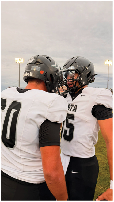 Two people wearing football helmets and white jerseys are standing close together on a field under a cloudy sky with stadium lights in the background. Description generated by AI