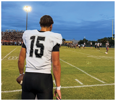 A person wearing a white sports jersey with the number 15 is standing on a football field watching a game in progress under a bright stadium light during dusk. Description generated by AI