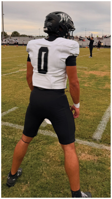 A person in a football uniform and helmet stands on a grassy field watching another person in black clothing throw a football with a small crowd in the bleachers under a cloudy sky. Description generated by AI