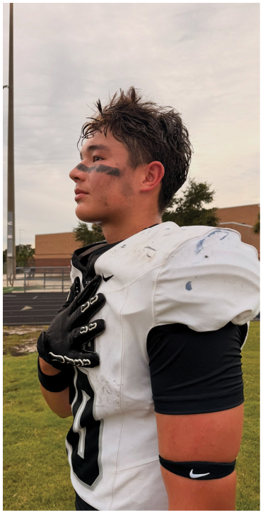A person wearing a white football uniform with black eye paint stands on a grassy field with one hand on their chest, looking to the side. Description generated by AI