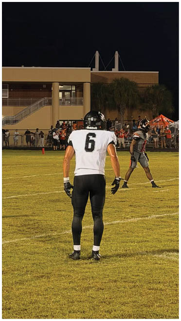 A person wearing a white sports jersey with the number 6 stands on a grassy field at night while other people and a referee are visible near a building in the background. Description generated by AI