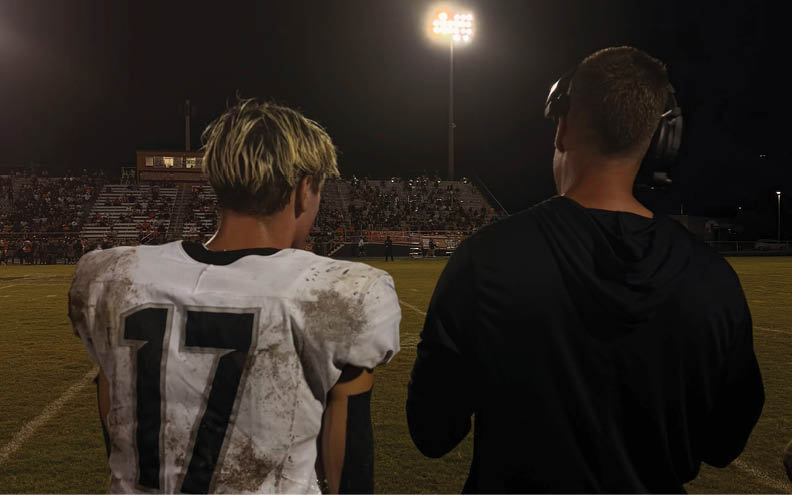 A person wearing a dirty football uniform stands next to another person wearing a headset and black clothing on a lit football field at night. Description generated by AI