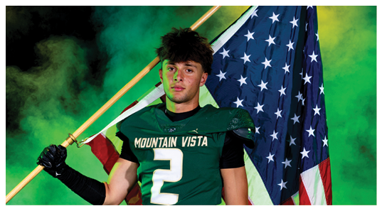 A person wearing a Mountain Vista sports jersey holds an American flag over their shoulder against a smoky green background. Description generated by AI