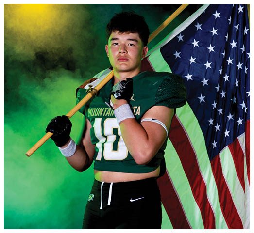 A person wearing a green football jersey and black pants holds a wooden pole over their shoulder with an American flag draped behind them against a green smoky background. Description generated by AI