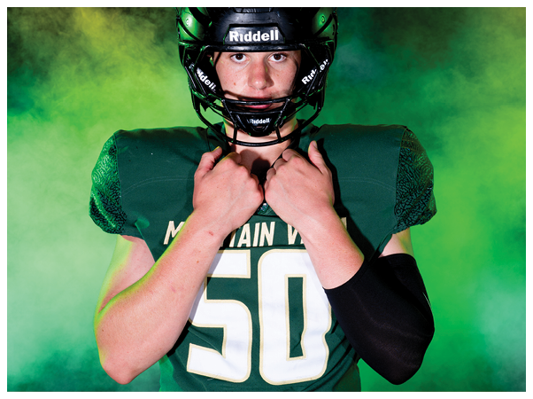 A person wearing a green football uniform and a black Riddell helmet is looking directly at the camera with a green and yellow smoky background. Description generated by AI
