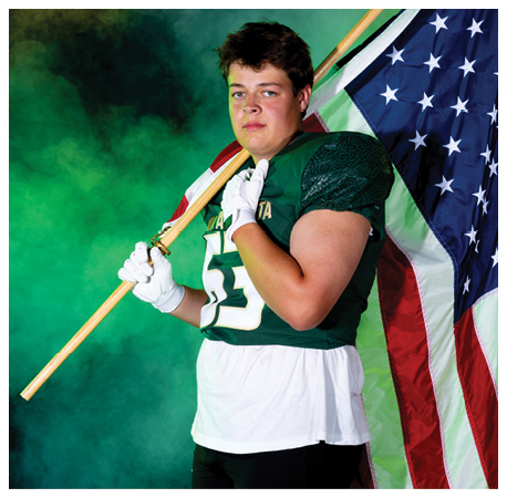 A person wearing a green sports jersey and white gloves holds an American flag over their shoulder against a green smoky background. Description generated by AI