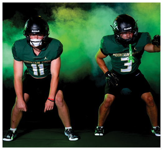 Two people in green football uniforms and black helmets are posed in a ready stance against a dark background with green smoke. Description generated by AI