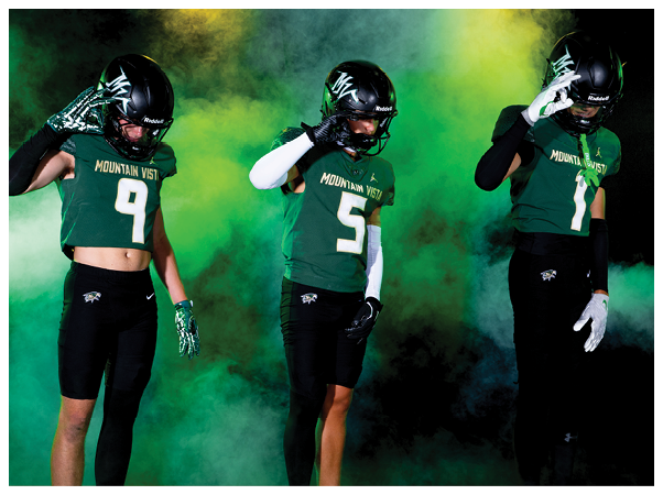 Three people wearing football helmets and green Mountain Vista jerseys stand in front of a colorful smoky background while making a hand gesture near their helmets. Description generated by AI