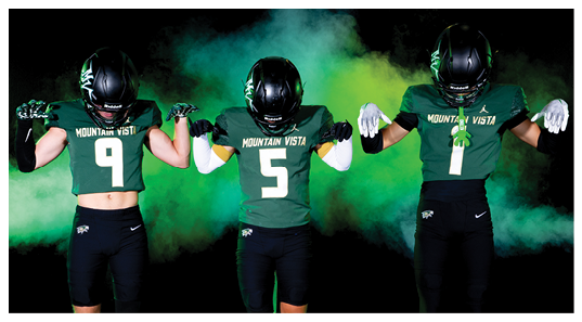 Three people wearing Mountain Vista football uniforms and helmets stand side by side against a smoky green and yellow background with their arms flexed to show strength. Description generated by AI
