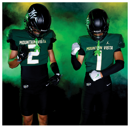 Two people wearing black and green football uniforms with helmets stand against a smoky green and yellow background, with one person looking down at their chest. Description generated by AI