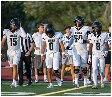Four people in football uniforms with helmets walk together on a field with a referee and others in the background. Description generated by AI