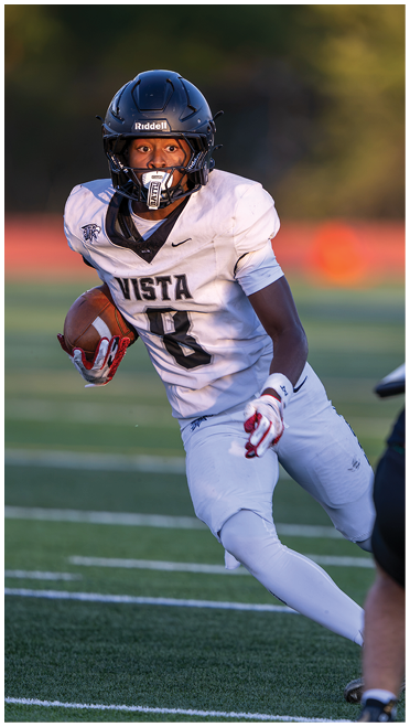 A person wearing a black helmet and white football uniform with the word Vista and number 8 is running on a football field while holding a football. Description generated by AI