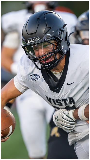 A person wearing a white football uniform with “VISTA" on the jersey is holding a football while another person in a dark uniform is grabbing their waist during a game. Description generated by AI