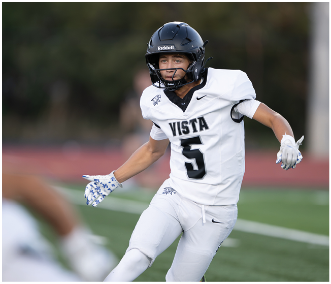 A person wearing a white football uniform with the word Vista and the number 5 is running on a field while wearing a black helmet and white gloves. Description generated by AI