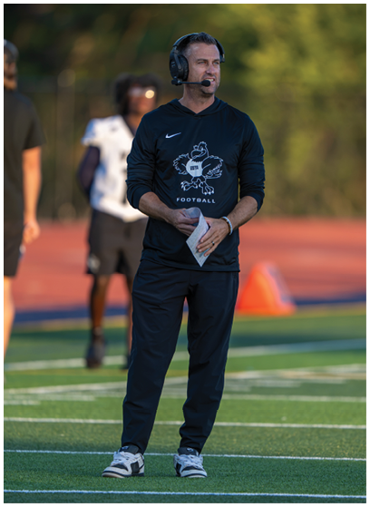 A person wearing a black Nike football hoodie and black pants stands on a sports field holding a folded piece of paper with another person blurred in the background. Description generated by AI