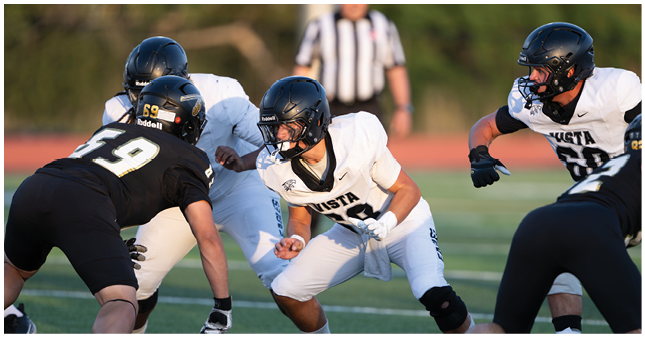 Two people in football uniforms and helmets are positioned facing each other on a field, appearing ready for a play. Description generated by AI