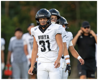 People wearing football helmets and uniforms are standing outdoors with a person in a black shirt and cap in the background. Description generated by AI