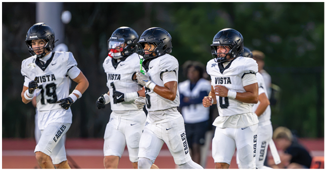 Four people in white football uniforms and black helmets are running on a field with a blurred background of other people and trees. Description generated by AI