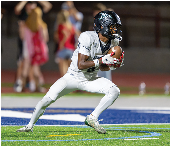 A person in a white football uniform and black helmet is crouched on a field holding a football, appearing ready to run or pass. Description generated by AI