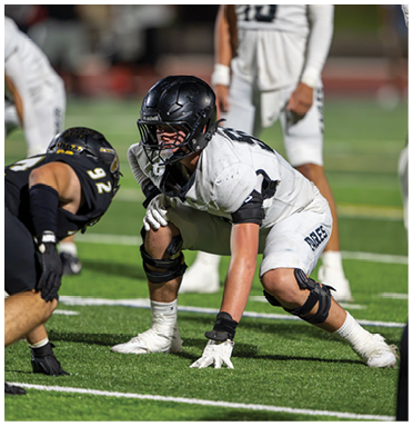 Two people in football uniforms and helmets are crouched facing each other on a green field, preparing for a play. Description generated by AI