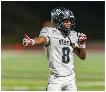 A person wearing a football uniform with the number 8 and the word Vista on the jersey is standing on a field, pointing with one hand and holding a mouthguard in the other. Description generated by AI