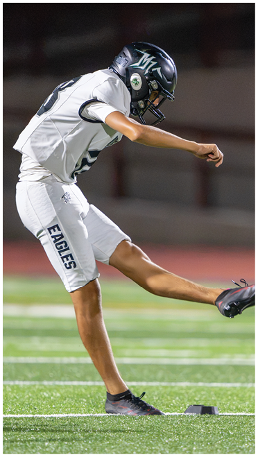 A person wearing a football uniform and helmet is kicking a football off a tee on a field. Description generated by AI