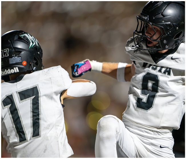 Two people in white football uniforms and black helmets are celebrating on the field during a game. Description generated by AI