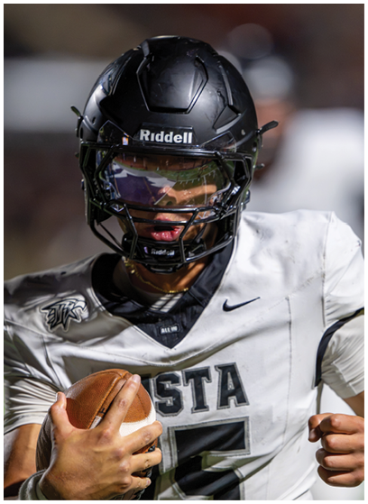 A person wearing a black football helmet and white jersey with the number 5 is holding a football and appears to be running or preparing to throw. Description generated by AI