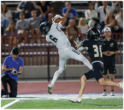 A person in a white sports uniform leaps to catch a football while another person in a black uniform attempts to block them on a lit football field with spectators in the background. Description generated by AI