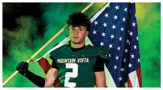 A person wearing a Mountain Vista sports jersey holds an American flag over their shoulder against a smoky green background. Description generated by AI