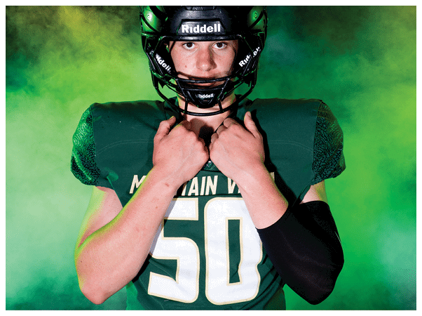 A person wearing a green football uniform and a black Riddell helmet is looking directly at the camera with a green and yellow smoky background. Description generated by AI