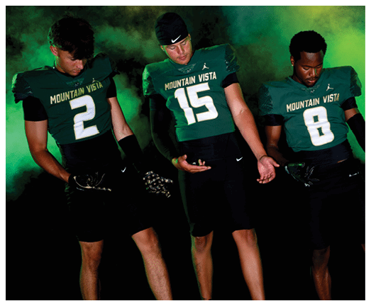 Three people wearing Mountain Vista football uniforms with numbers 2, 15, and 8 stand against a smoky green and yellow background, looking down at their hands. Description generated by AI