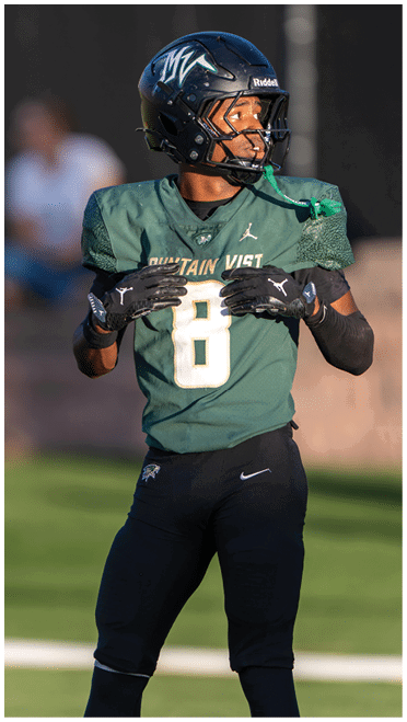 A person wearing a blue football helmet and green jersey is shown from the side against a blurred dark background. Description generated by AI