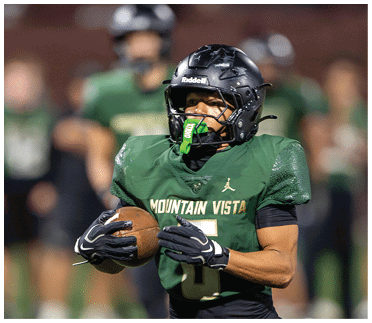 A person wearing a black football helmet and green Mountain Vista jersey is holding a football with blurred people in green jerseys in the background. Description generated by AI